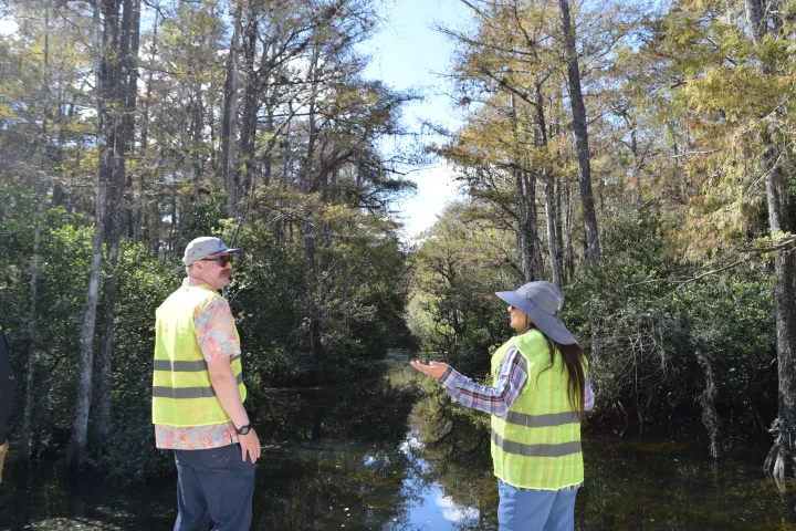 Two people in vests and hats talk in a forest with water and tall trees around them.